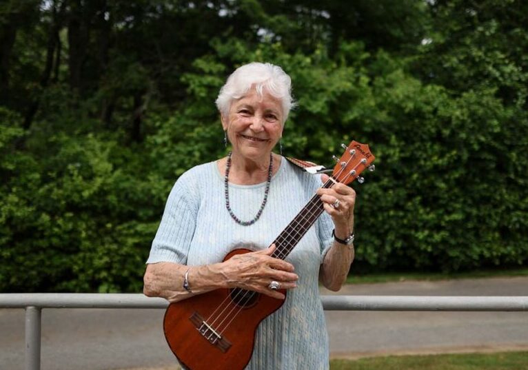 Older adult woman smiling outdoors while holding and playing a ukulele, standing in front of greenery and a paved path, conveying joy and engagement in music.