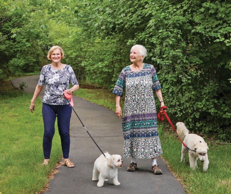 Two residents walk side by side along a paved path surrounded by lush greenery, each holding a leash as they stroll with their dogs on a scenic campus walking trail.