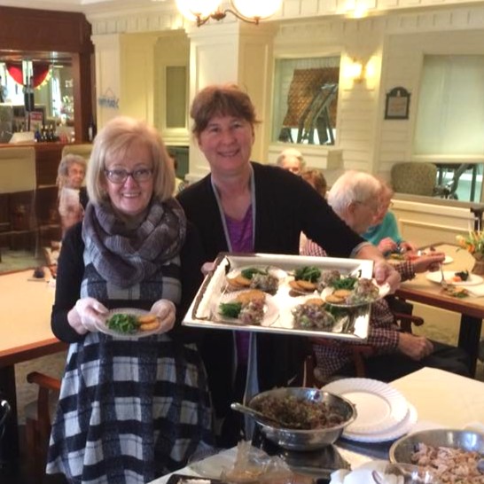 Two smiling women presenting a tray of prepared food in a dining area. The woman on the left wears glasses and a black-and-white plaid dress with a gray scarf, holding a small plate of food. The woman on the right wears a black cardigan over a purple top and holds a larger tray with several plated servings. In the background, other residents sit at tables, engaged in conversation.