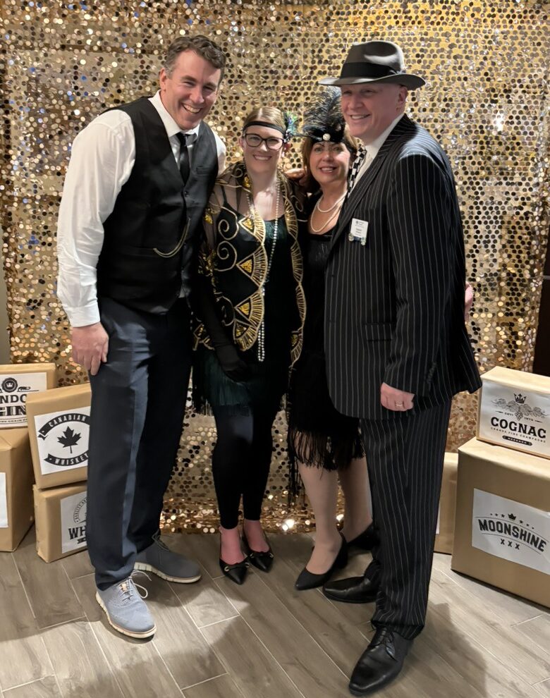 Four adults dressed in Roaring ’20s–themed attire pose together in front of a gold sequin backdrop, smiling for a photo during a festive celebration.