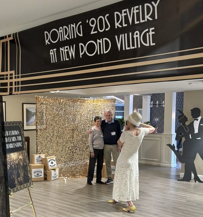 Guests pose for a photo in front of a gold sequin backdrop beneath a “Roaring ’20s Revelry at New Pond Village” banner, while a woman in a vintage-style dress and hat takes their picture in a festively decorated community space.
