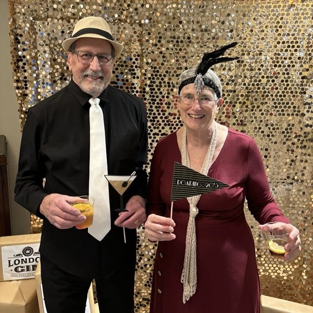 Two smiling residents dressed in 1920s attire stand in front of a gold sequin backdrop, holding cocktails and a “Roaring ’20s” party prop—he wears a black shirt with a white tie and fedora, and she wears a burgundy flapper-style dress with a beaded headpiece.