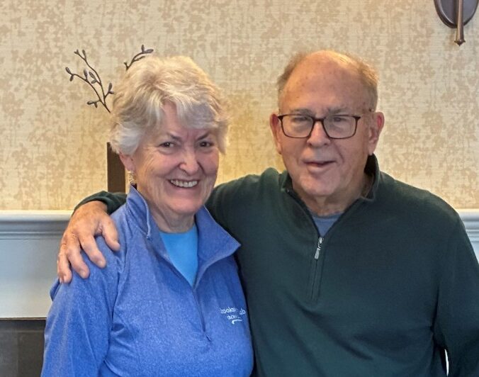 An older couple stands smiling with their arms around each other in front of a fireplace. The woman wears a blue pullover and black pants, and the man wears glasses, a green quarter-zip sweater, and khaki pants. They appear relaxed and happy in a warmly lit indoor setting.