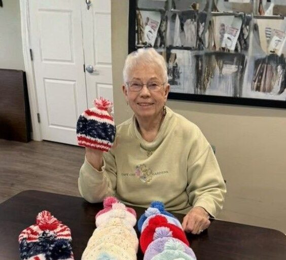 Shirley Hutchins sits at a table smiling and holding a hand-knit winter hat, with several colorful knit hats and blankets laid out in front of her.