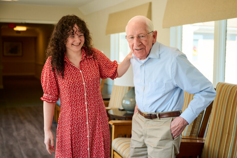 A woman in a red dress smiles beside an older man in a blue shirt as they walk together through a bright, welcoming community space.