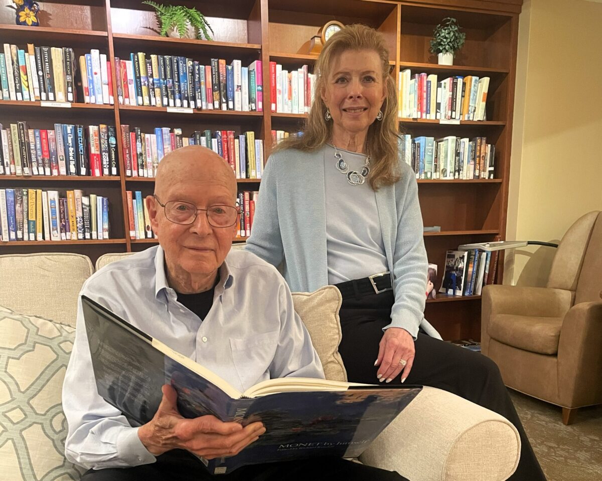 New Pond Village residents Jerry and Leslie Reicher pose together in the community library as Jerry looks through a large art book.